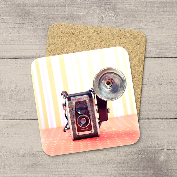 A square cork coaster with rounded corners featuring a printed vintage double-lens Kodak camera on a striped yellow-and-white background, placed on a gray wood plank surface.