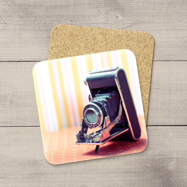 A square coaster with rounded corners featuring a print of a Sears Tower vintage camera, displayed on a cork backing over a gray wood plank surface.