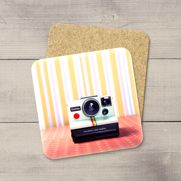 A square printed coaster with rounded corners lies on a cork backing over a light wood surface. The coaster design shows a retro Polaroid instant camera on a pink-orange floor against a pastel striped background.