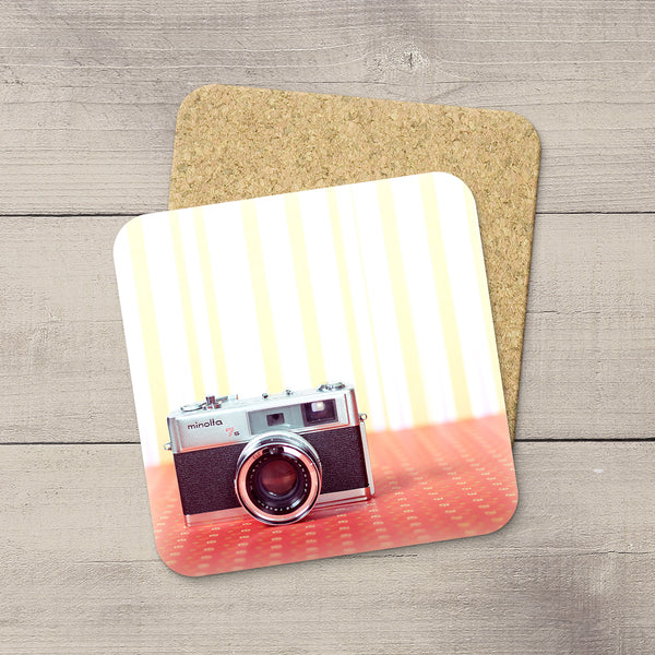 A square cork coaster with rounded corners featuring a printed design of a vintage camera on a striped cream and red background, displayed on a gray wood plank surface.