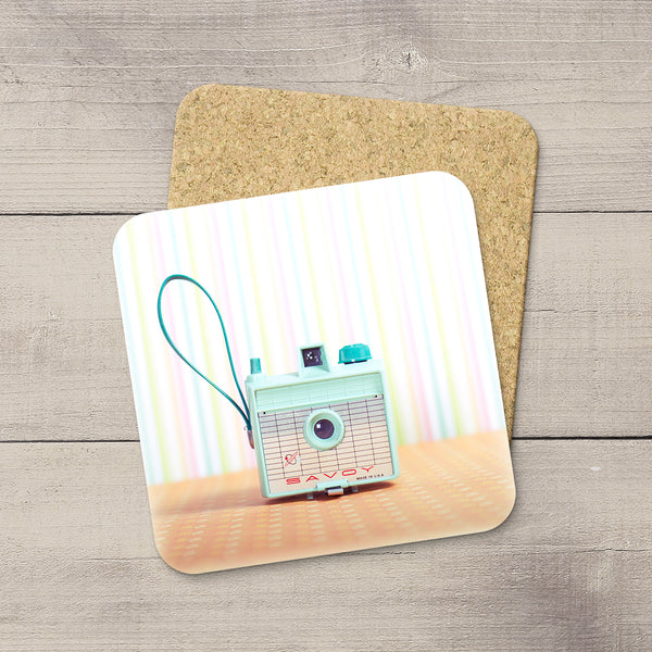 A square printed coaster with rounded corners lies on a light wood surface, partially covering a cork backing. The printed design shows a retro camera in mint green and turquoise tones against pastel vertical stripes and a peach-colored tabletop.