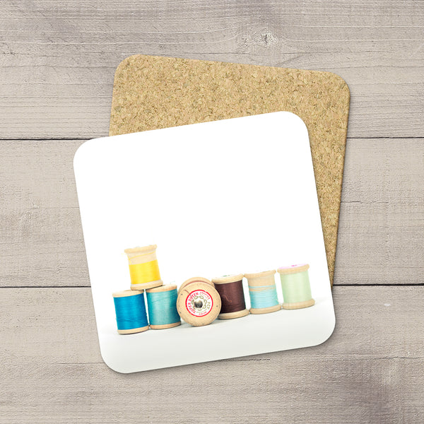 A square white coaster with rounded corners featuring a printed design of colorful sewing thread spools, displayed on top of a cork coaster over a gray wood plank background.