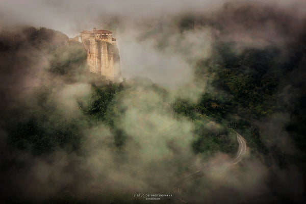 Misty aerial landscape photograph featuring a tall stone tower or cliff structure rising above dense green forest, with a winding road visible through the fog. The scene has a moody, atmospheric composition with low light and heavy haze.