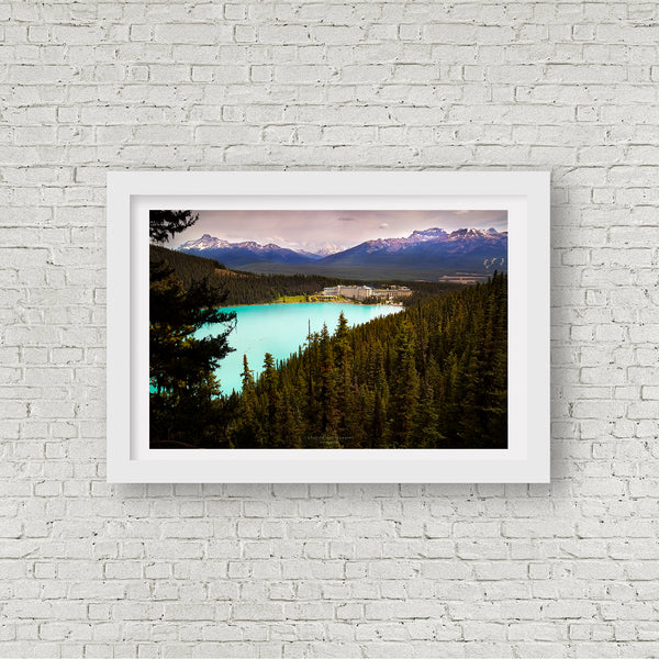 A framed landscape photograph of Lake Louise and the Chateau surrounded by dark pine forests and distant snow-capped mountains, displayed on a white brick wall.