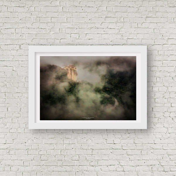 Framed print of a Byzantine monastery on a Greek cliff in Meteora, surrounded by dense morning mist, mounted on a white brick wall.