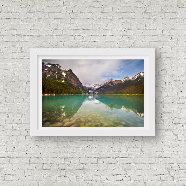 A framed landscape photograph of a turquoise alpine lake reflecting snow-capped mountains and a blue sky, displayed on a white brick wall.