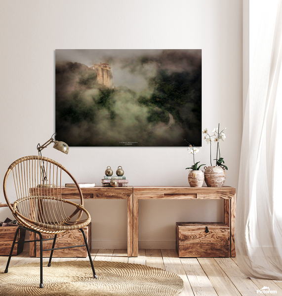 A large rectangular wall art print depicting a misty mountain landscape hangs above a wooden console table in a styled room with a wicker chair, lamp, vases, and white flowers. Meteora Monastery.