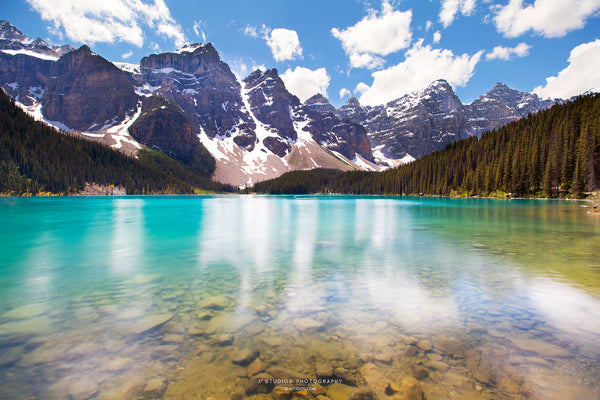 A scenic landscape photograph of a turquoise alpine Moraine Lake in Alberta with steep snow-dusted mountains, dark pine forests, and a bright blue sky with white clouds reflecting on the water surface.