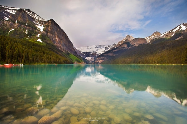 A scenic landscape photograph of a turquoise Lake Louise with snow-capped mountains, evergreen forests, and dramatic clouds reflected in the clear water.