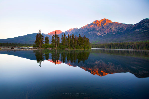 A scenic landscape photograph of a mountain range reflected in a calm lake, with a small island of evergreen trees and mountains lit by warm orange sunlight on the horizon.