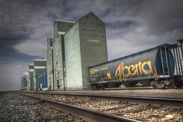 Archival picture of grain elevator row in Warner. An Alberta hopper train car on the train tracks is also featured.
