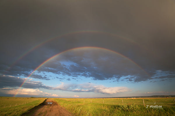 Beautiful image of a double rainbow arcing over an inviting country road and a farmers field filled with bales of hay. Canadian Photography.